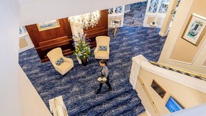 A hotel lobby with a blue carpet, two armchairs, a table with a vase of flowers, a chandelier, and a staircase.