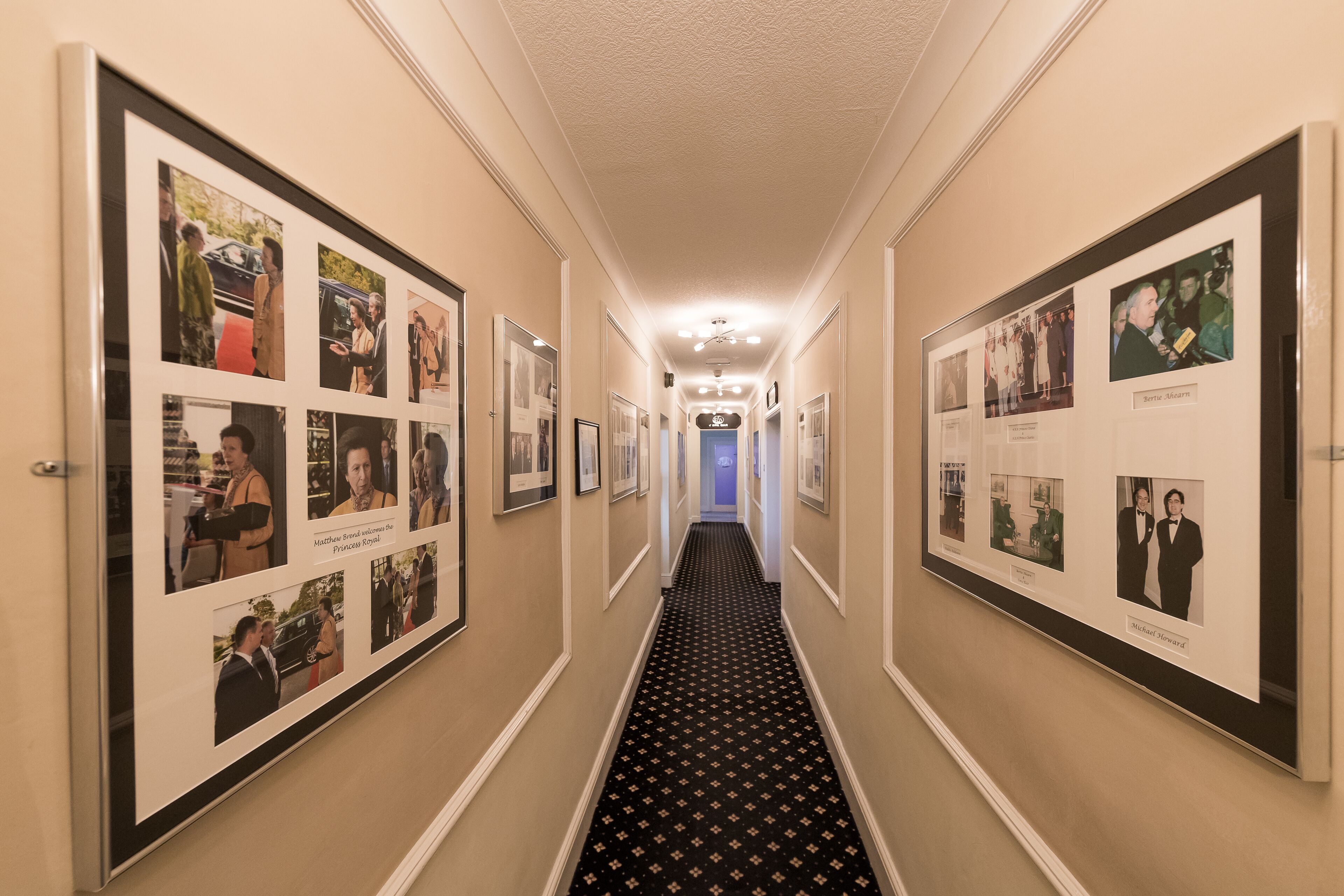 A hallway with framed photographs on the walls, a patterned carpet, and a ceiling light fixture.