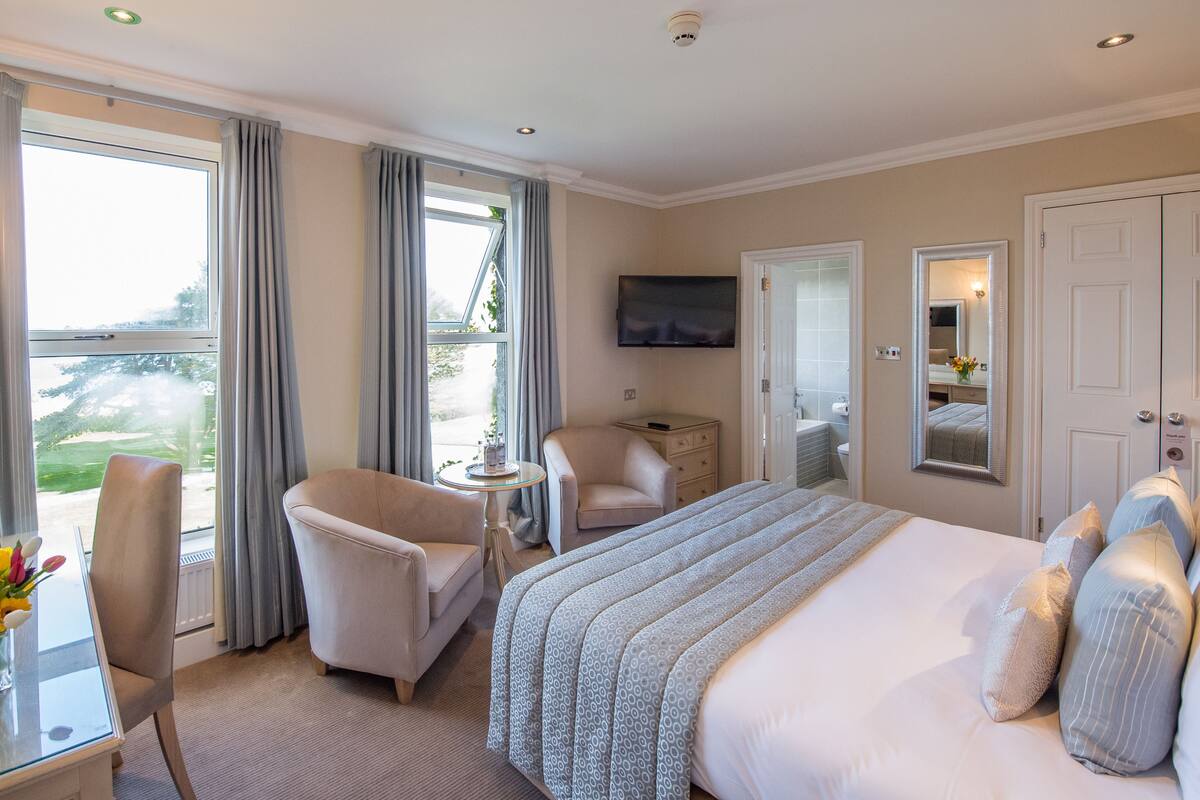 A hotel room with a large bed, a sitting area with a chair and a table, a TV mounted on the wall, and a view of the outdoors through the window.