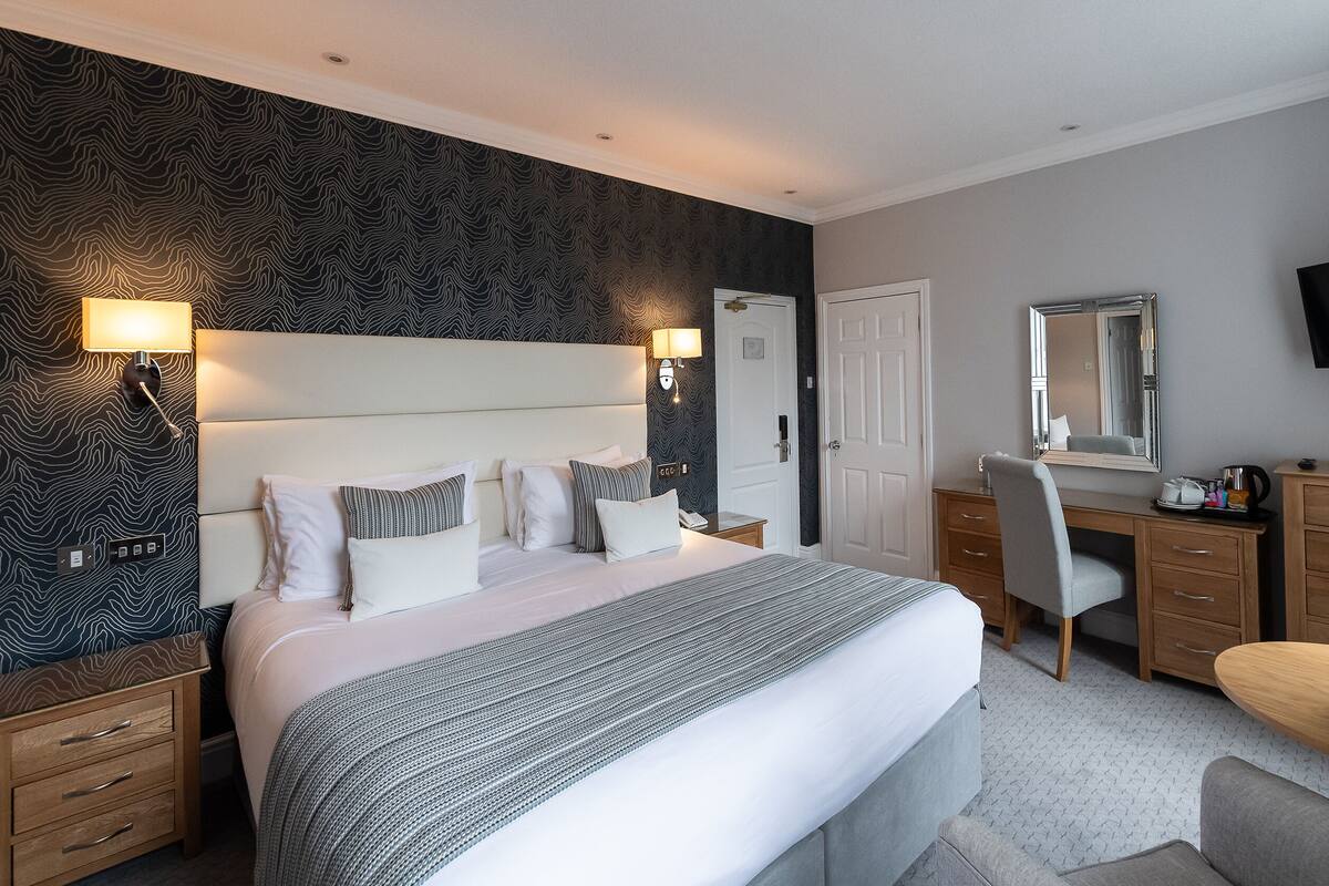 A hotel room with a large bed, bedside tables, a desk, a chair, a TV, and a mirror.