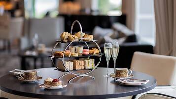 A three-tiered dessert stand with various pastries and sandwiches, two glasses of champagne, and a cup of coffee on a round table in a hotel setting.