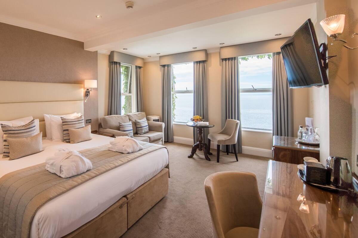 A hotel room with a large bed, a desk, a chair, a TV, and a view of the sea.