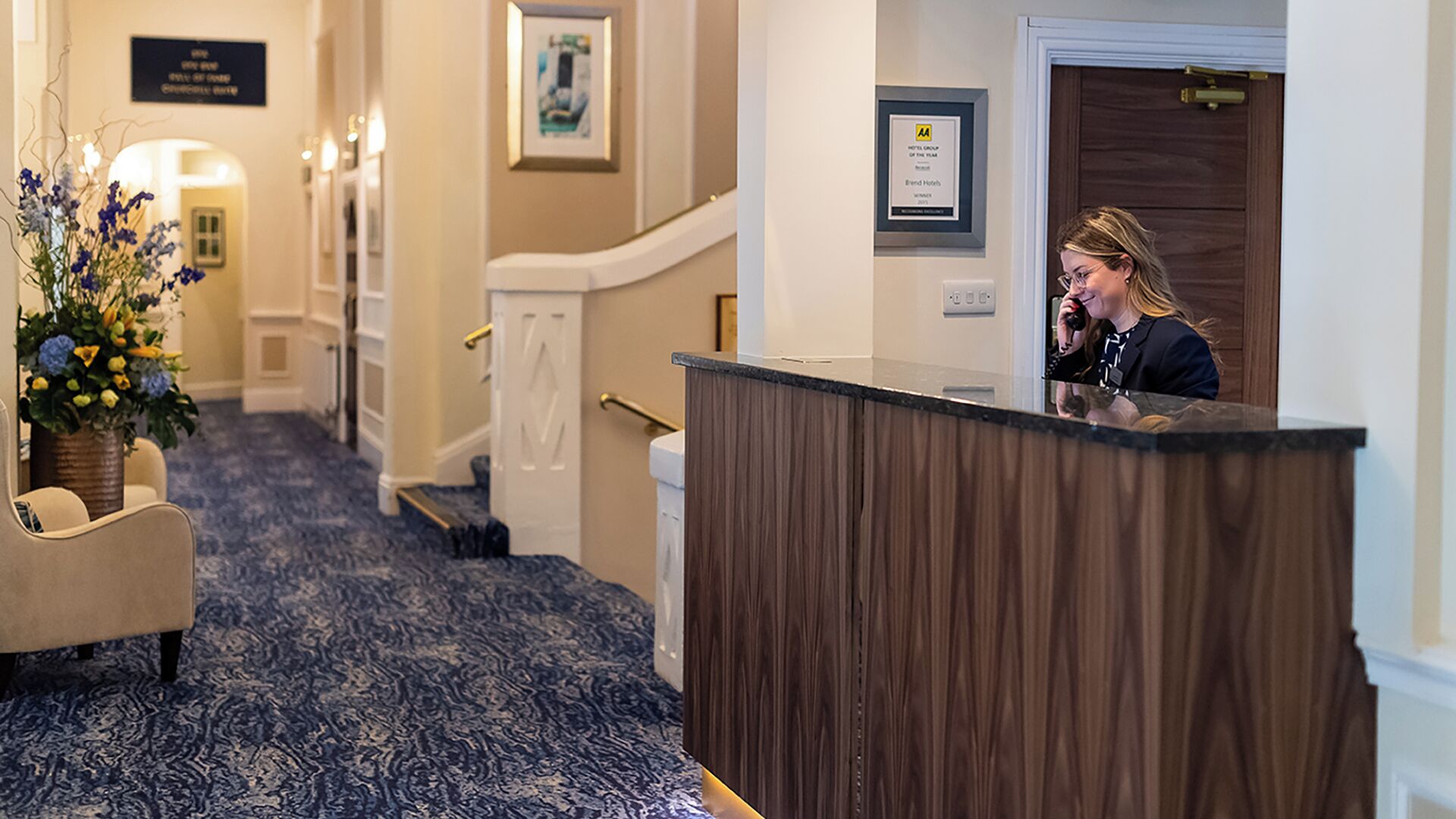 A hotel reception area with a reception desk, a staircase, and a hallway with a patterned carpet.
