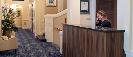 A hotel reception area with a reception desk, a staircase, and a hallway with a patterned carpet.