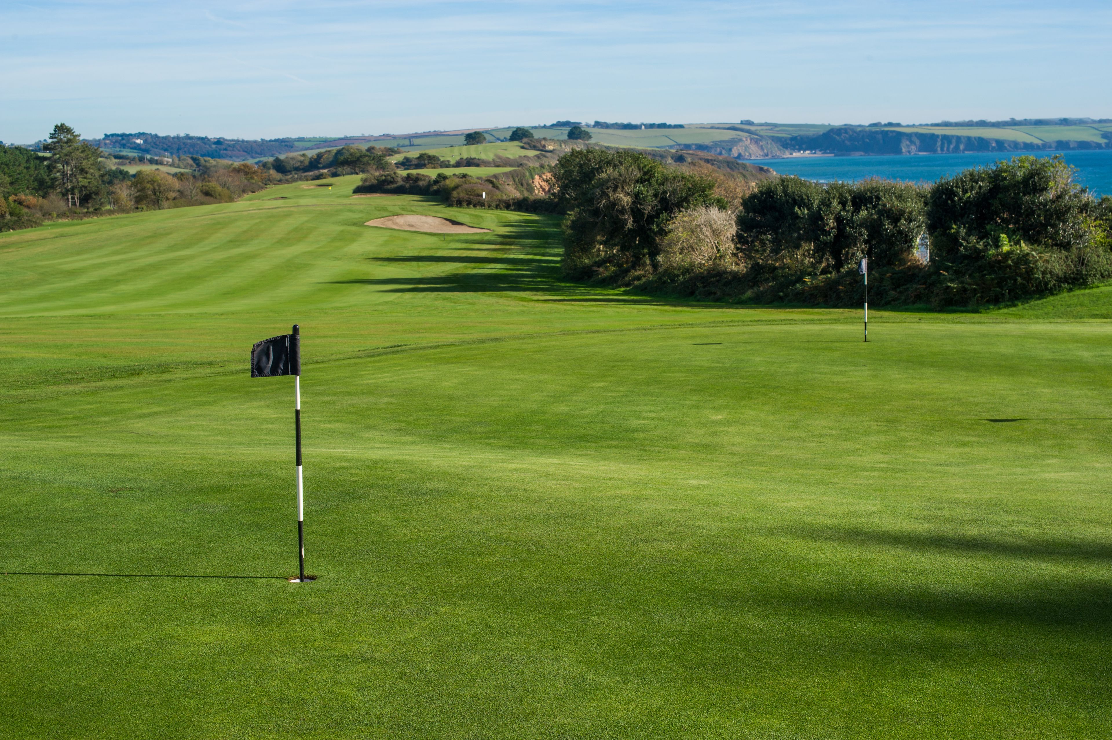Un campo de golf con un mástil de bandera, césped cuidado y vista al mar.