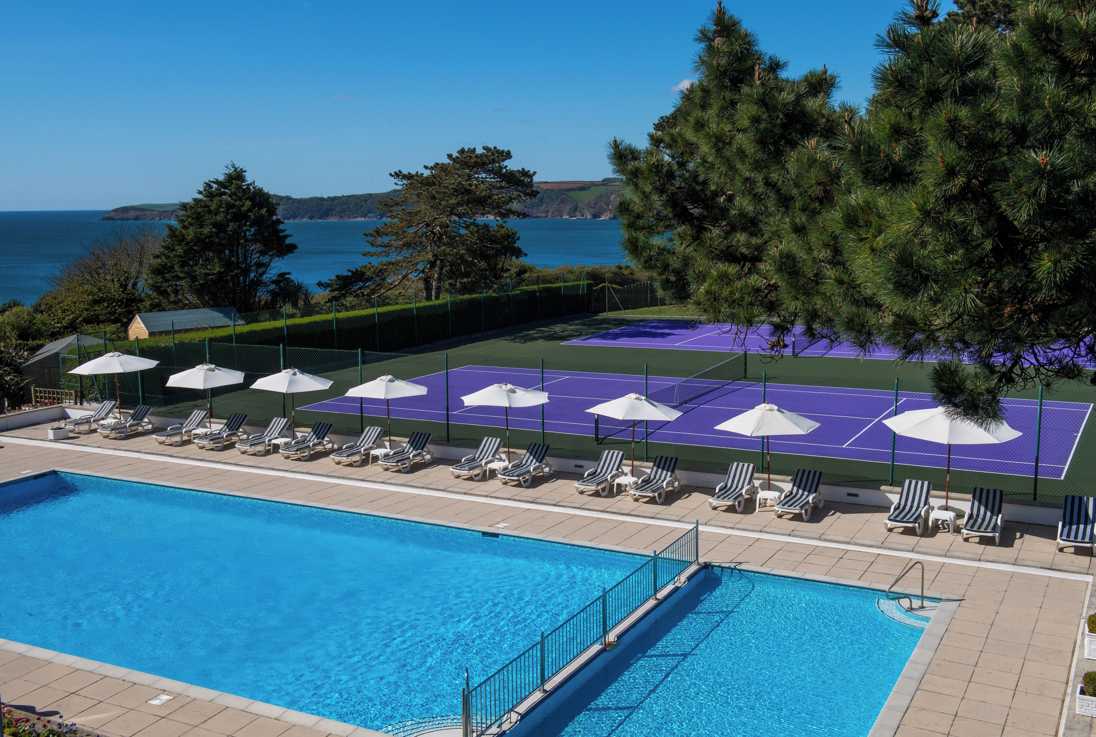 Un área de piscina con cancha de tenis, sillas de descanso y sombrillas.