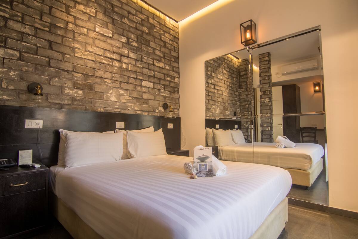 A hotel room with a brick wall, a large bed, a bedside table with a phone and a menu, and a mirror reflecting the room.