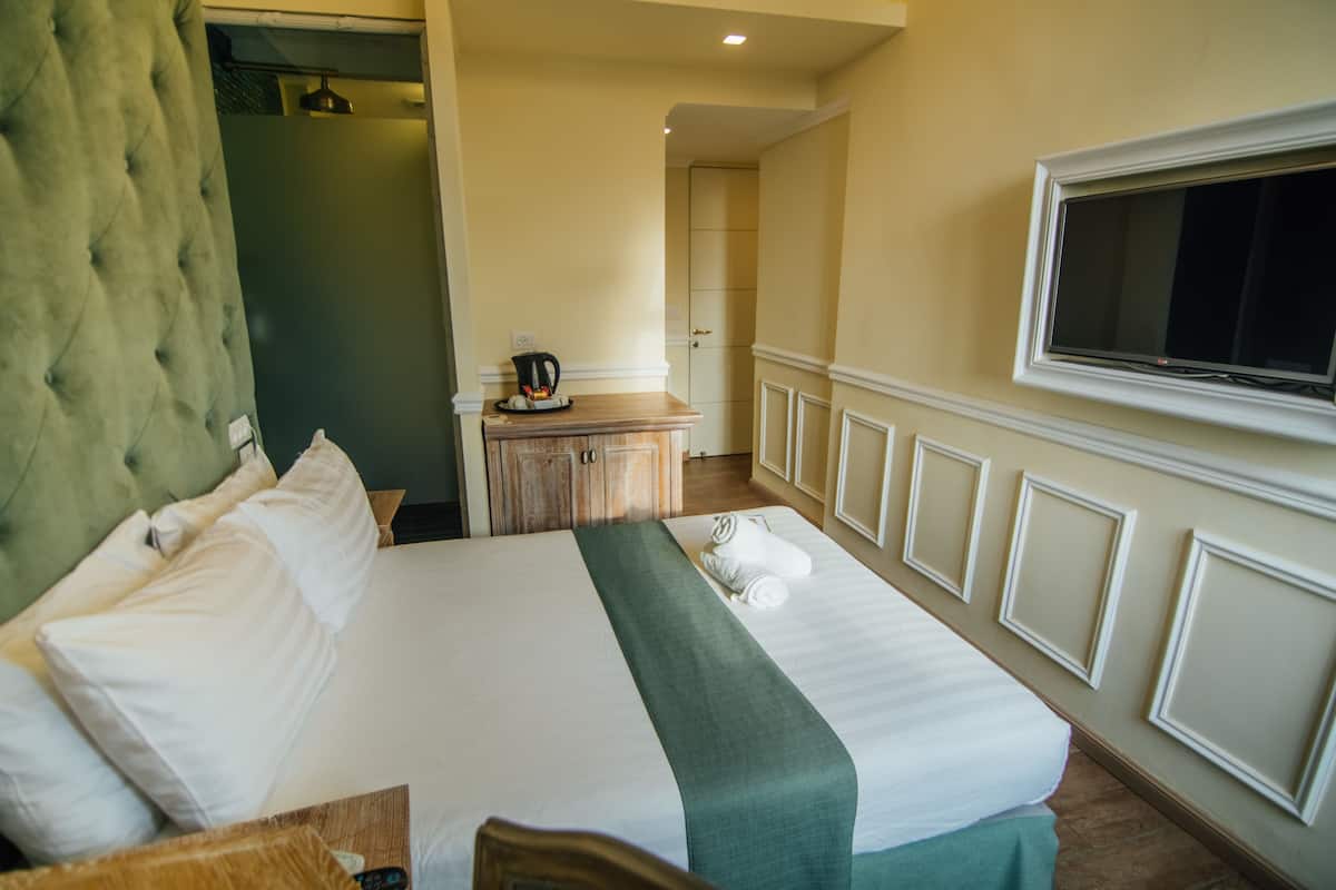 A hotel room with a large bed, a wooden dresser, a television mounted on the wall, and a green upholstered headboard.