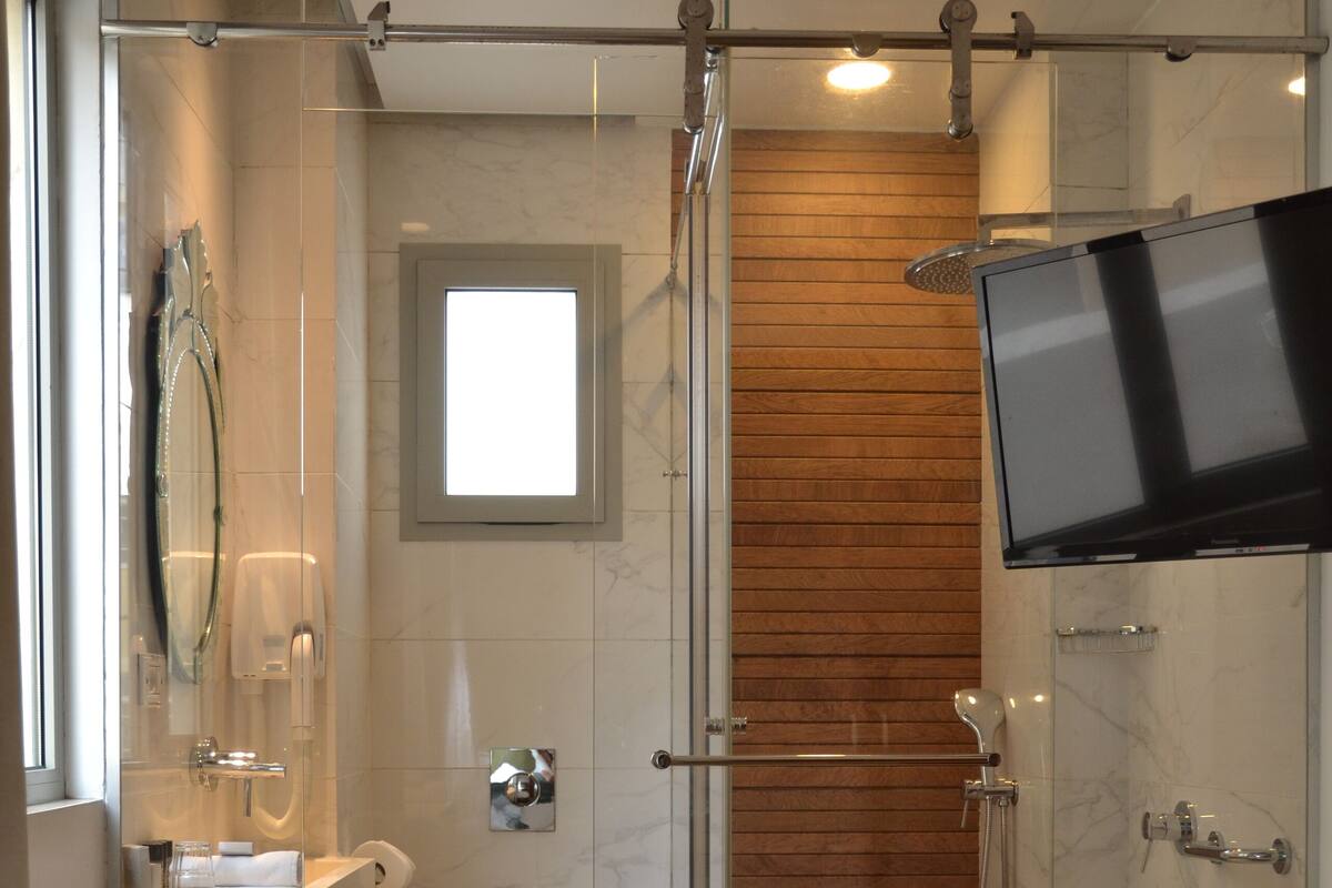A modern bathroom with a glass-enclosed shower, a wooden vanity, a toilet, and a flat-screen TV mounted on the wall.