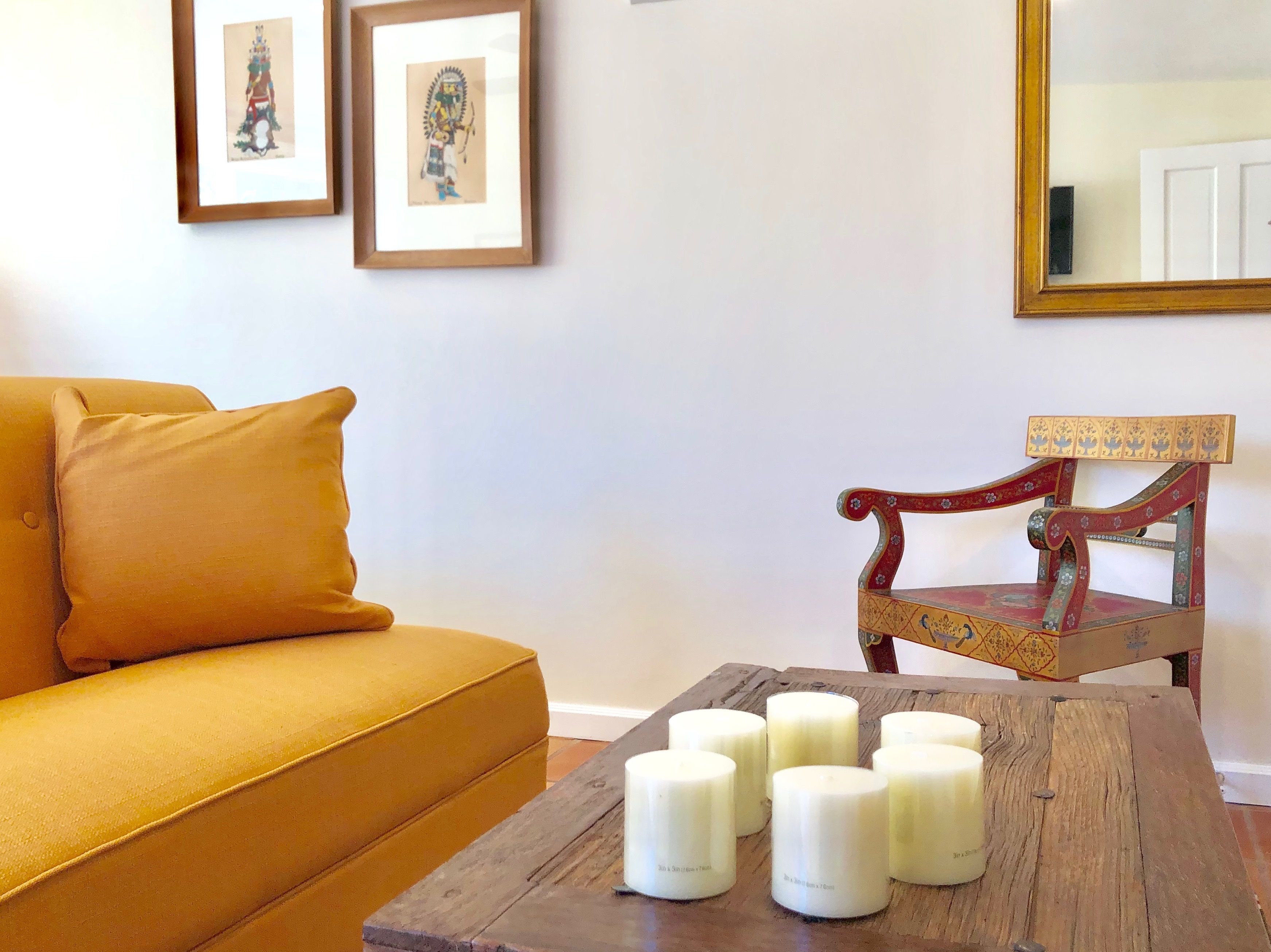 Una sala de estar con un sofá color mostaza, una mesa de centro de madera con velas, una silla de madera y dos cuadros en la pared.