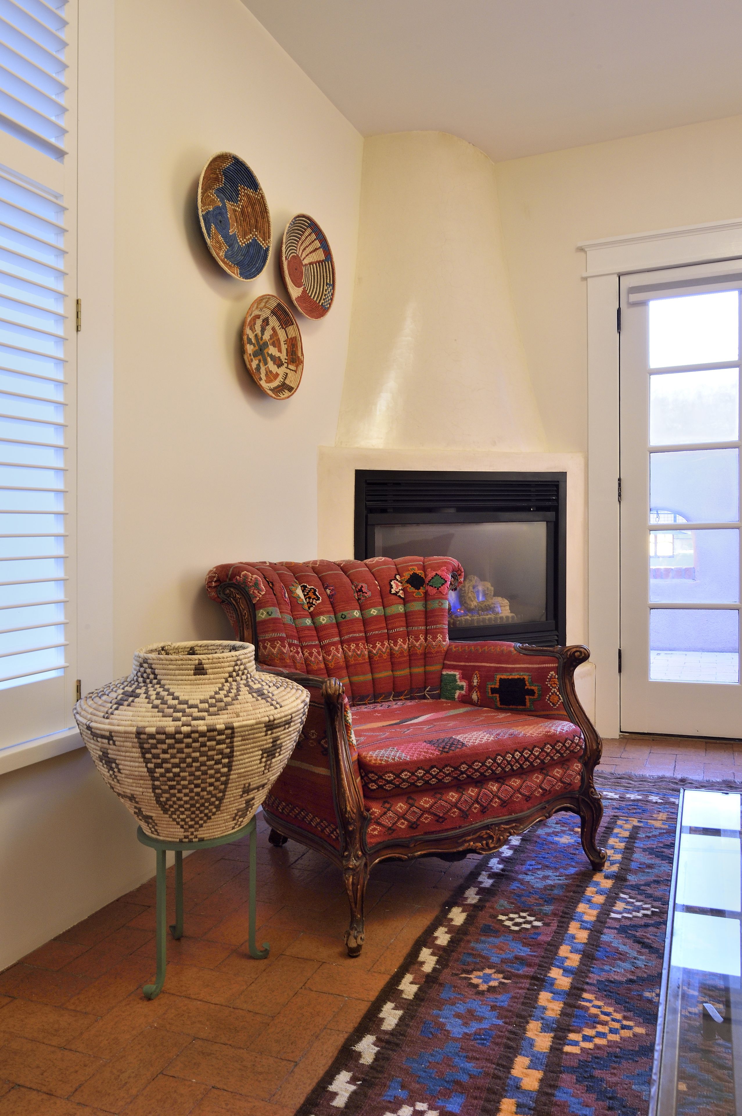 A living room with a fireplace, a patterned armchair, a woven basket, and decorative plates on the wall.