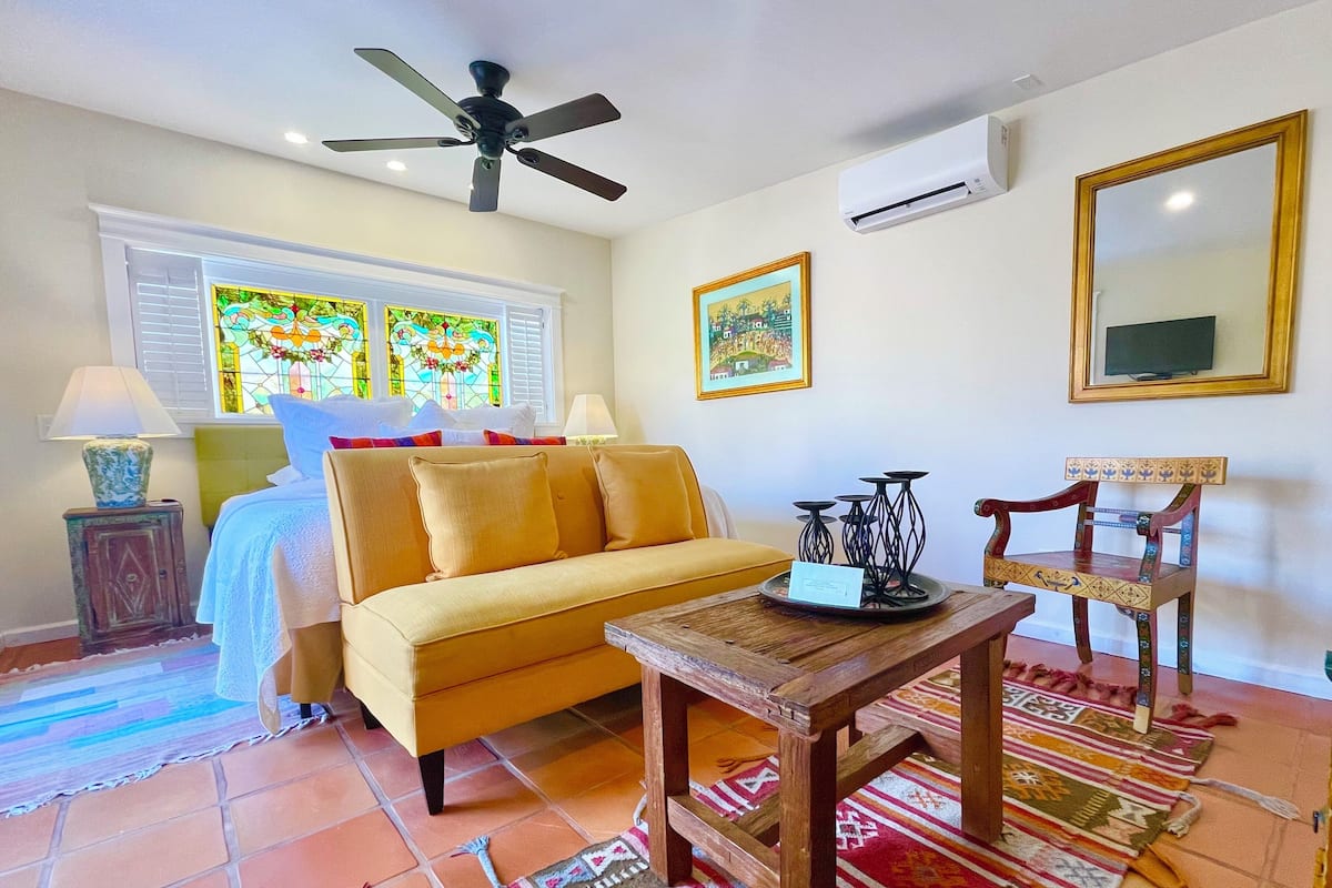 A cozy room with a yellow sofa, a wooden coffee table, a decorative chair, a ceiling fan, and a stained glass window.