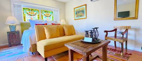 Una habitación acogedora con un sofá amarillo, una mesa de centro de madera, una silla decorativa, un ventilador de techo y una ventana con vidrieras.