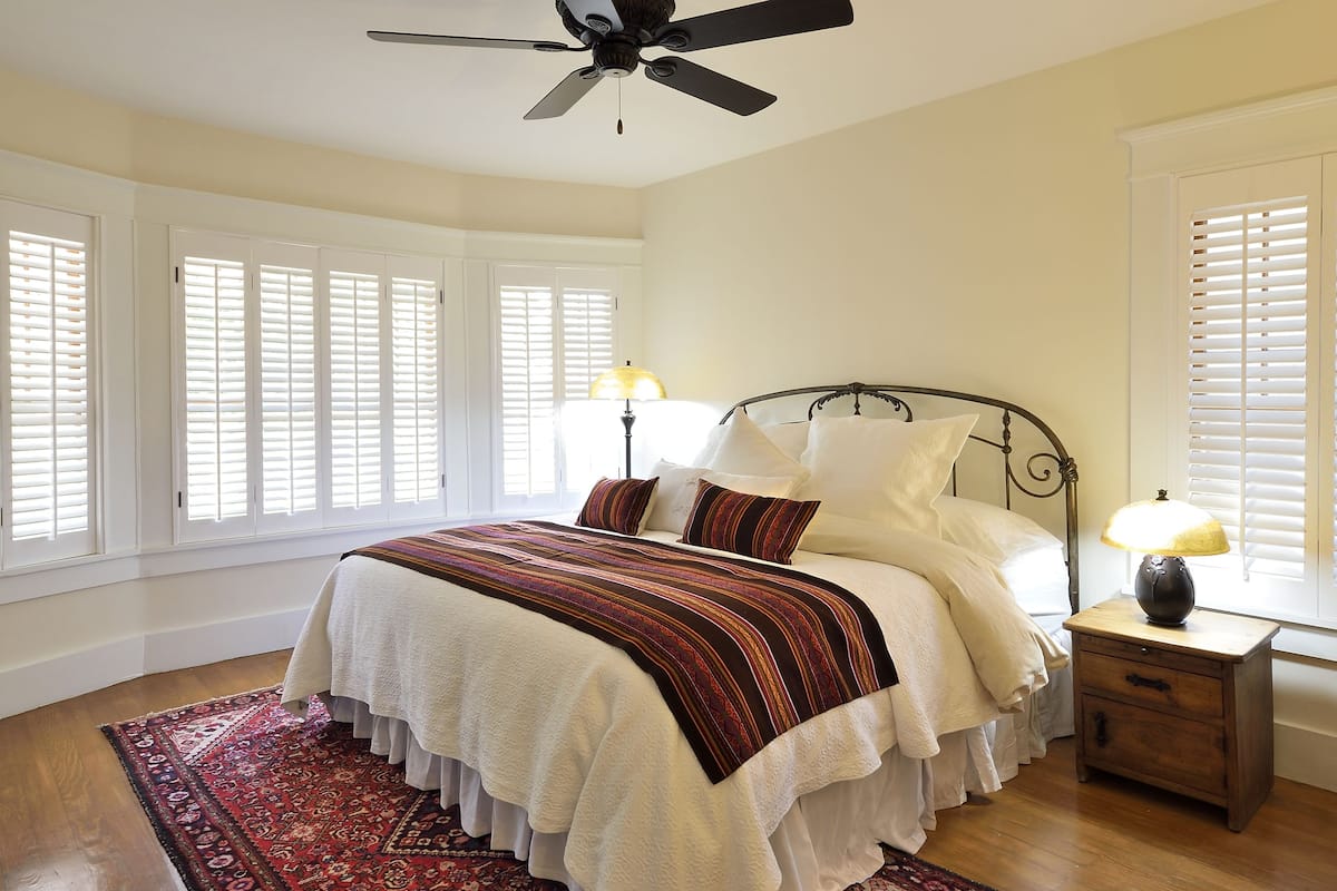 A bedroom with a bed, a nightstand, a lamp, a ceiling fan, and a window with blinds.