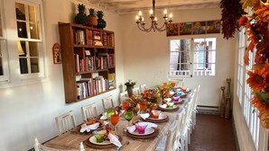 Un comedor con una larga mesa de madera dispuesta para una comida, rodeada de sillas blancas. Se ve una estantería llena de libros a la izquierda.