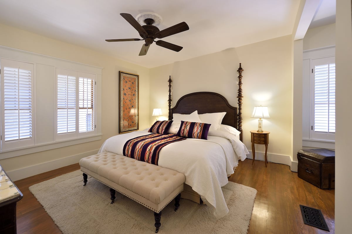 A bedroom with a large bed, a ceiling fan, a bench, and a nightstand with a lamp.