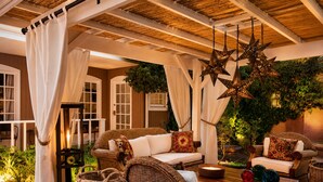 Un área de estar al aire libre techada con mobiliario de mimbre, una mesa con esferas de vidrio decorativas y luces colgantes en forma de estrella.