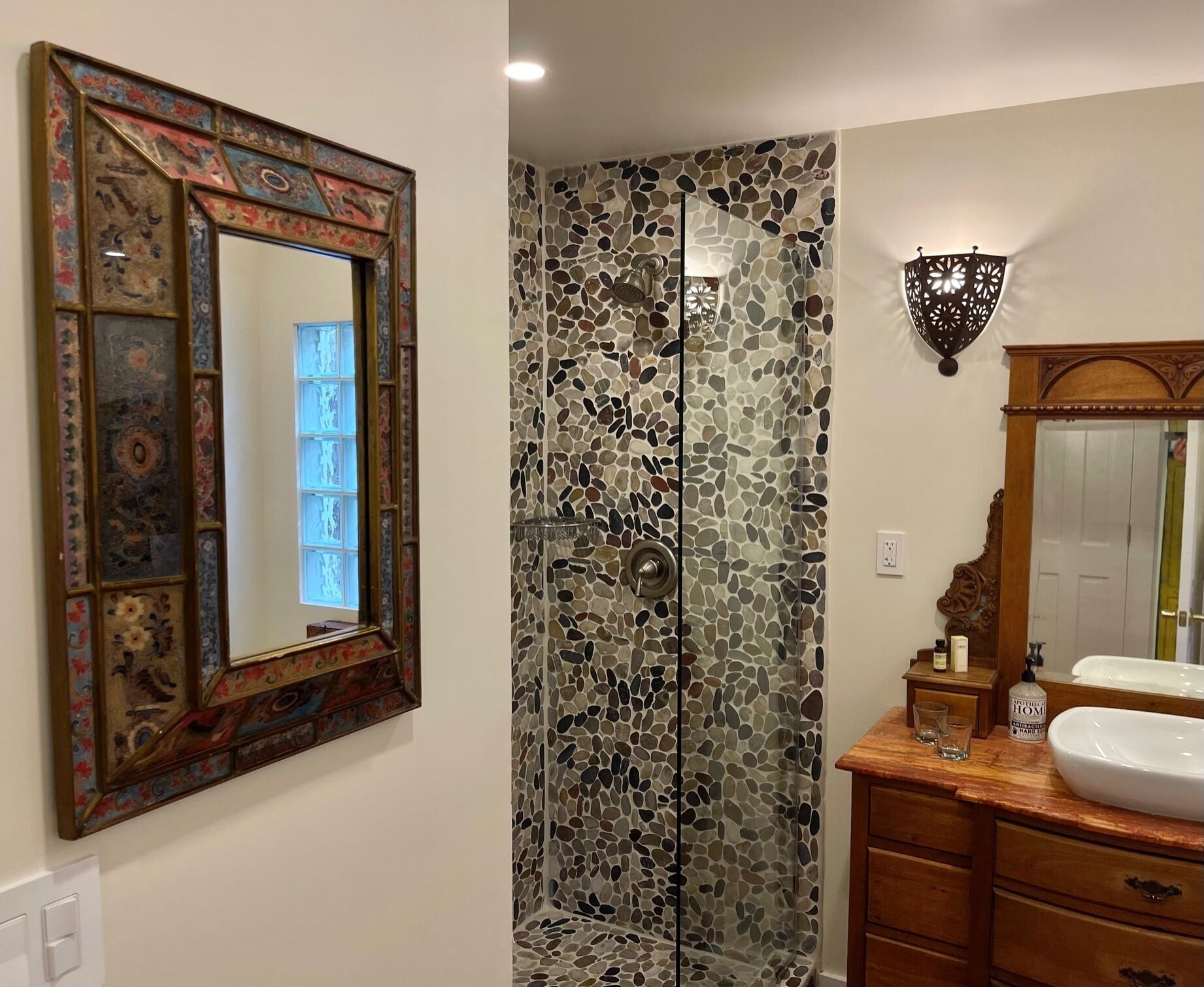 A bathroom with a mosaic shower wall, a wooden vanity with a white sink, and a decorative mirror with a colorful frame.