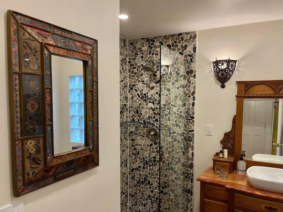 A bathroom with a mosaic shower wall, a wooden vanity with a white sink, and a decorative mirror with a colorful frame.