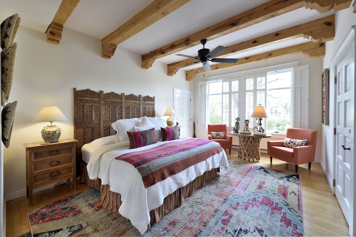 A bedroom with a large bed, wooden ceiling beams, a ceiling fan, a window with shutters, a small table with a lamp, and a chair.
