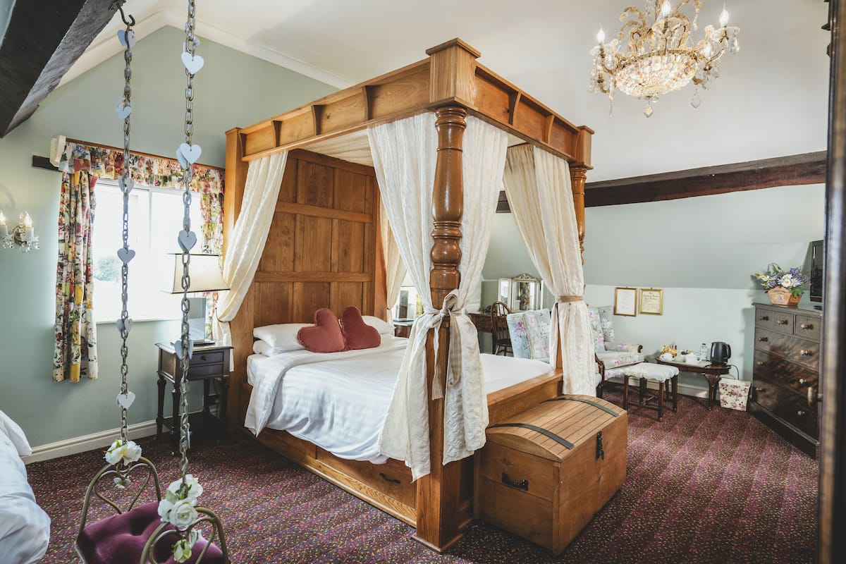 A four-poster canopy bed with a wooden bench and chest of drawers, a chandelier, and a floral window dress.