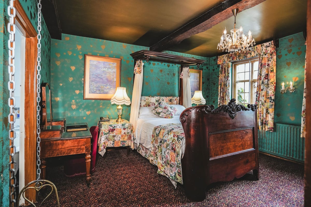 A room with a four-poster bed, a wooden desk, a chair, a nightstand with a lamp, and a chandelier.