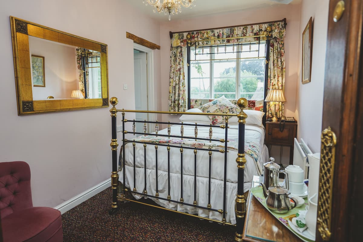 A bedroom with a four-poster bed, a large mirror, floral curtains, and a view of greenery outside.