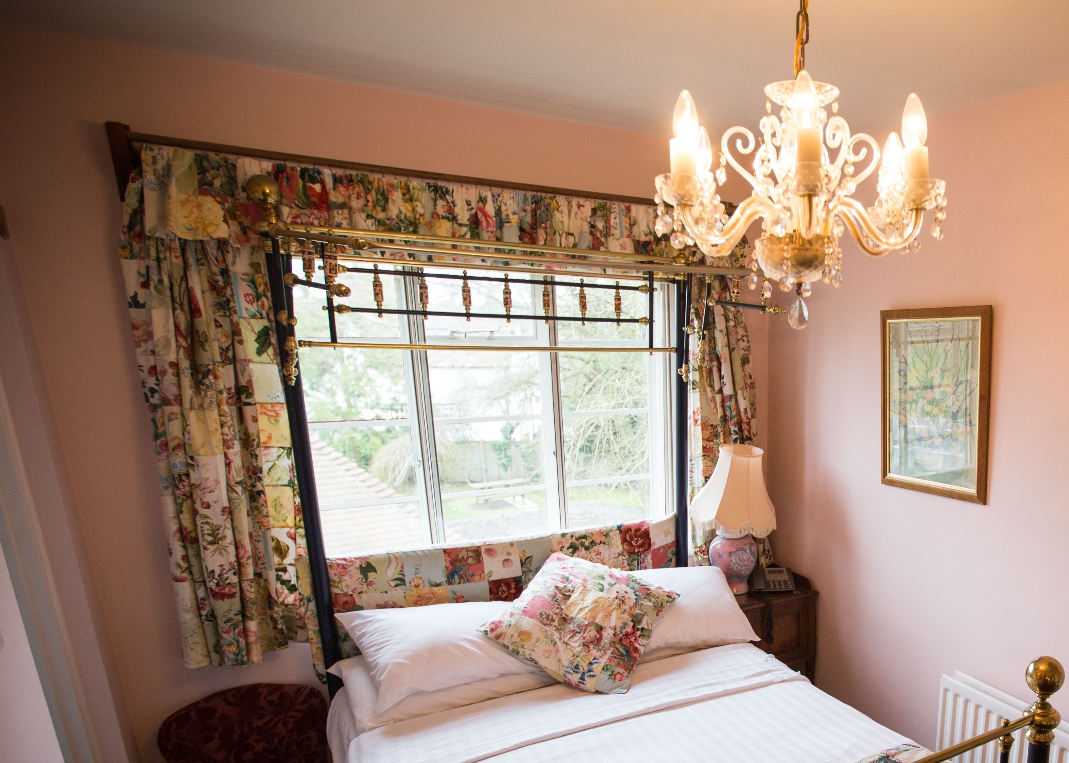 A bedroom with a bed, floral curtains, a chandelier, a bedside table with a lamp, and a framed picture on the wall.