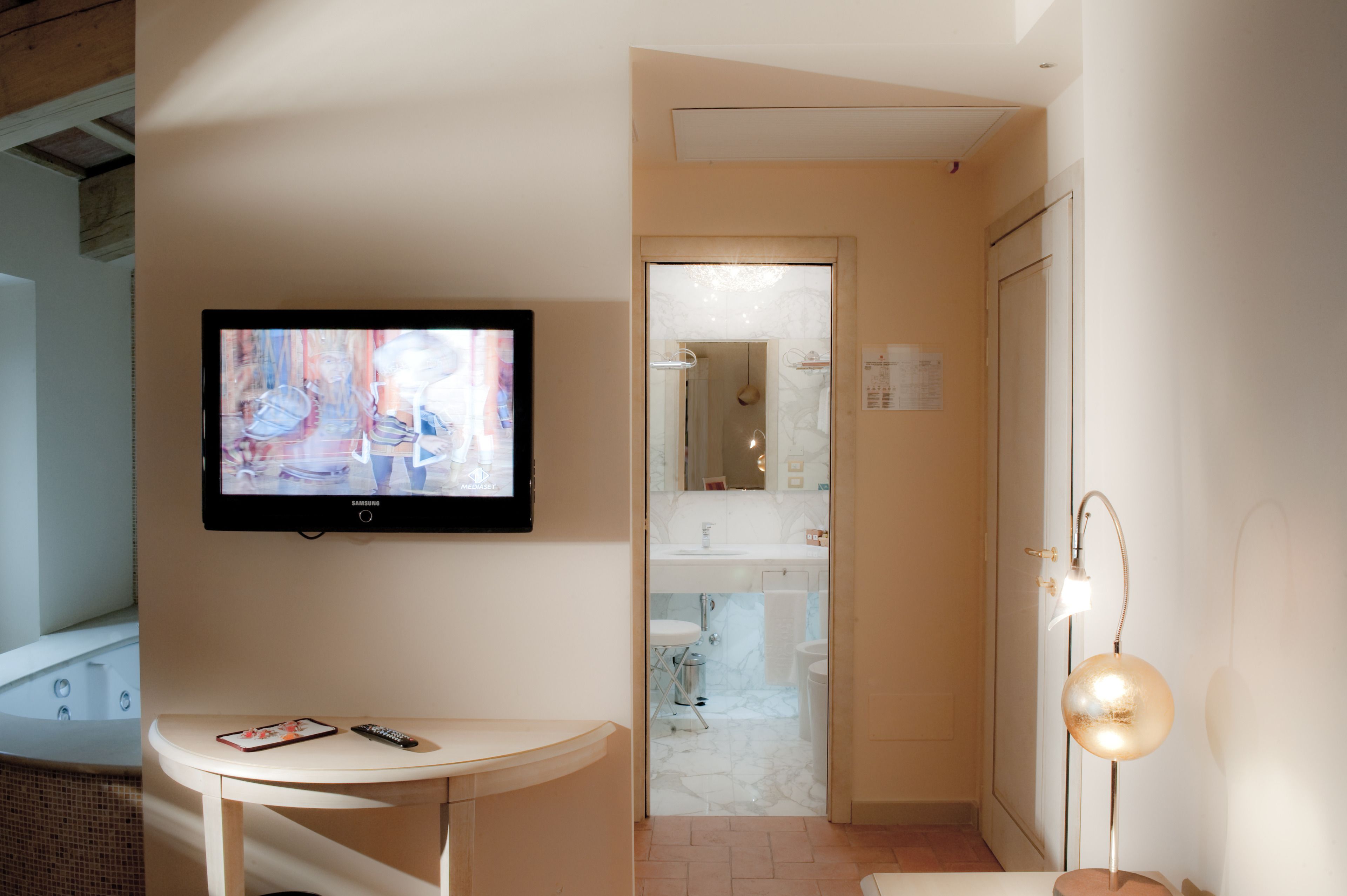 Un baño moderno con una televisión montada en la pared, una mesita con un control remoto y un teléfono, y una lámpara de pie.