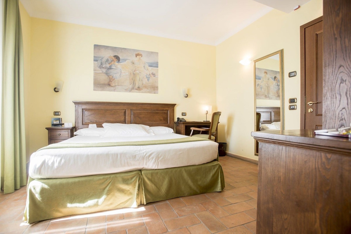 A hotel room with a large bed, bedside tables, a desk, a chair, a mirror, and a painting on the wall.