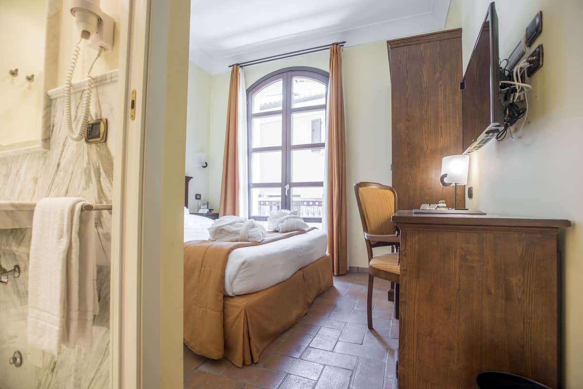 A hotel room with a bed, a wooden desk, a chair, a window with curtains, and a telephone.
