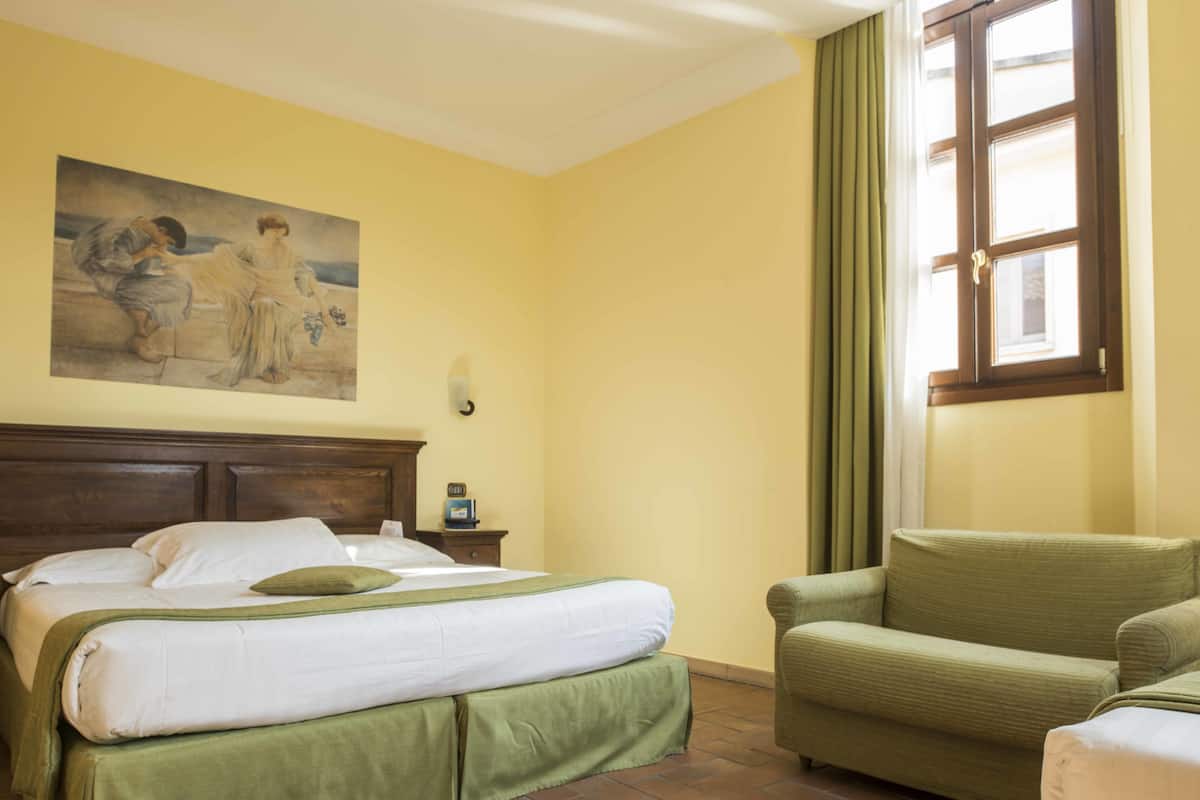 A hotel room with a bed, a green armchair, a painting on the wall, and a window with curtains.