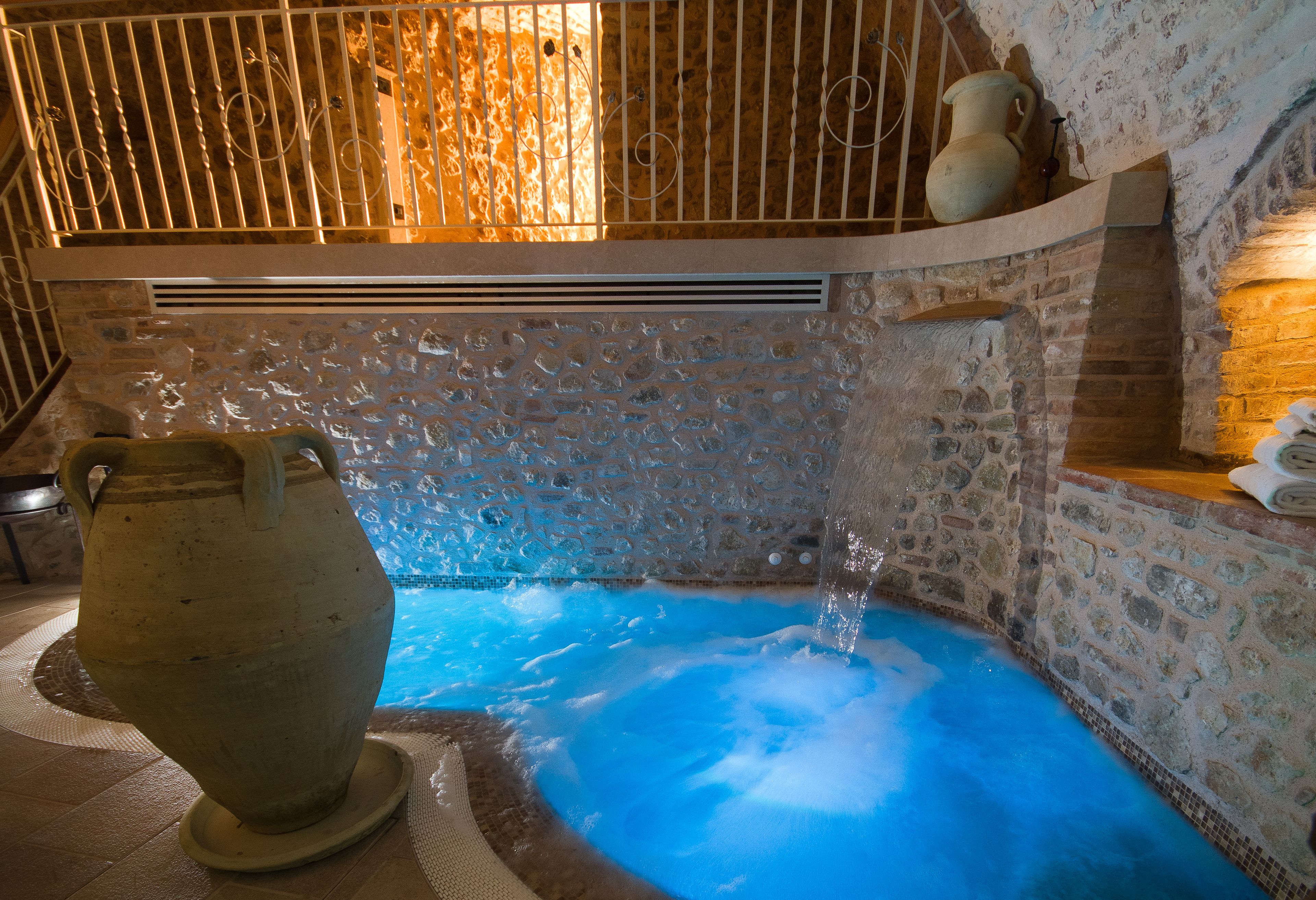 Een hottub met stenen muren, een helderblauw wateroppervlak, een grote keramieken pot en een houten plank met decoratieve objecten.