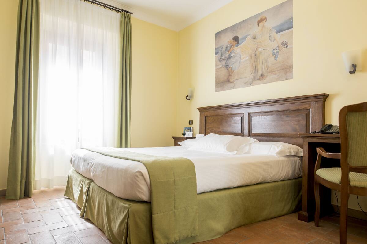A hotel room with a bed, a chair, a nightstand, a painting on the wall, and a window with curtains.
