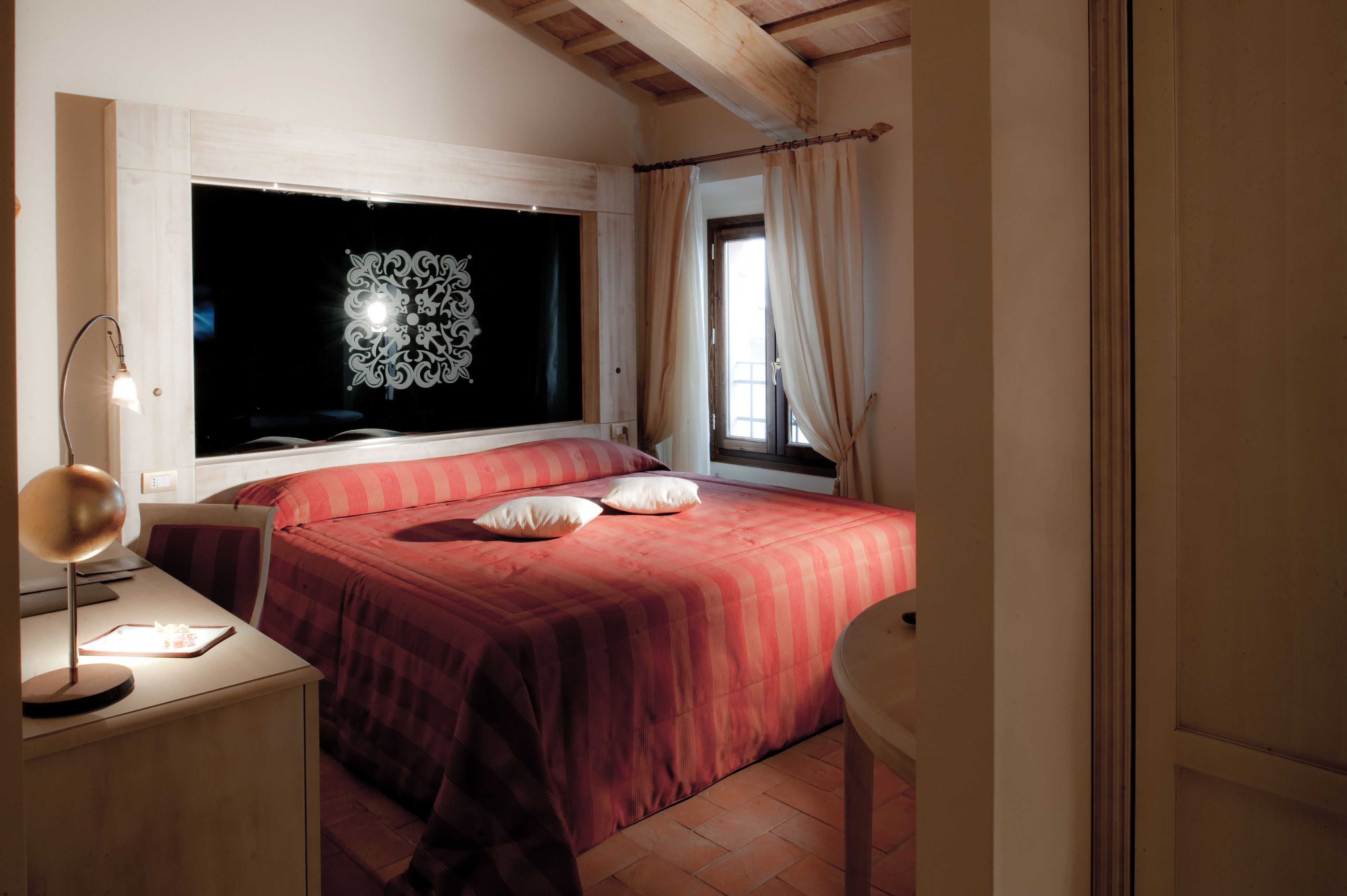 Un dormitorio con un ventanal grande, una cama con ropa de cama roja, una mesita de noche con una lámpara y una silla.