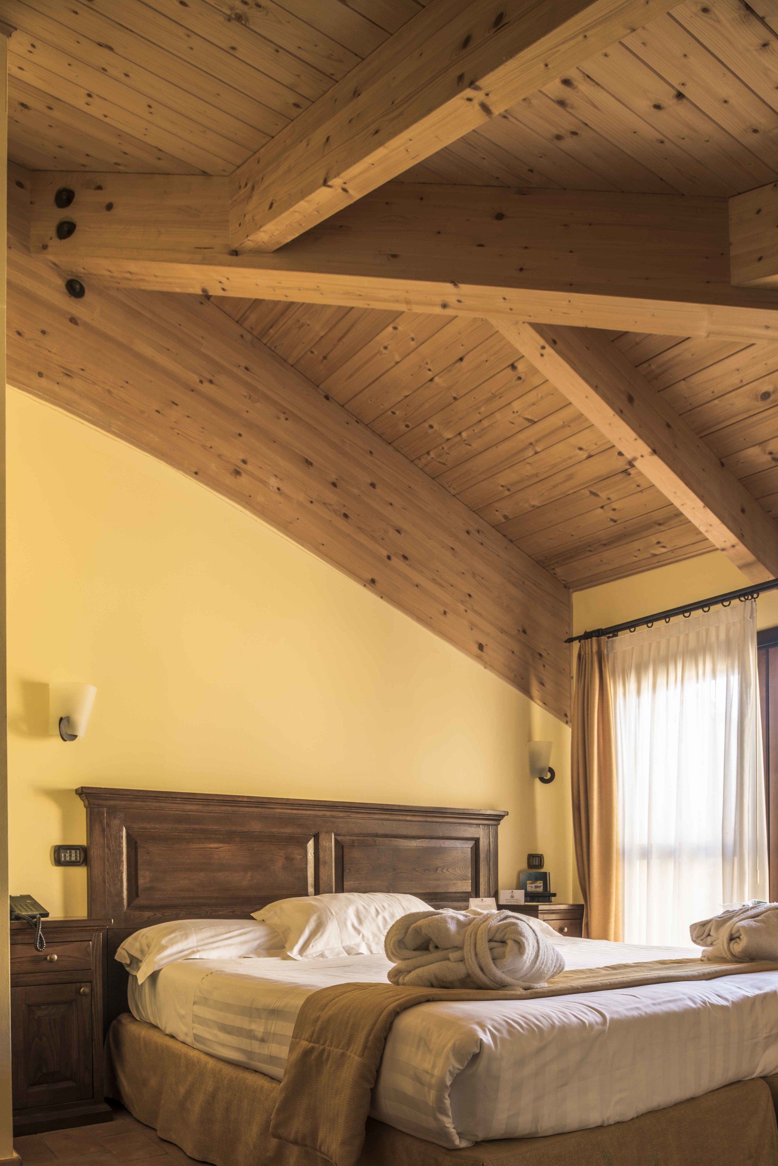A bedroom with a wooden ceiling, a bed with white linens, a nightstand, and a window with curtains.