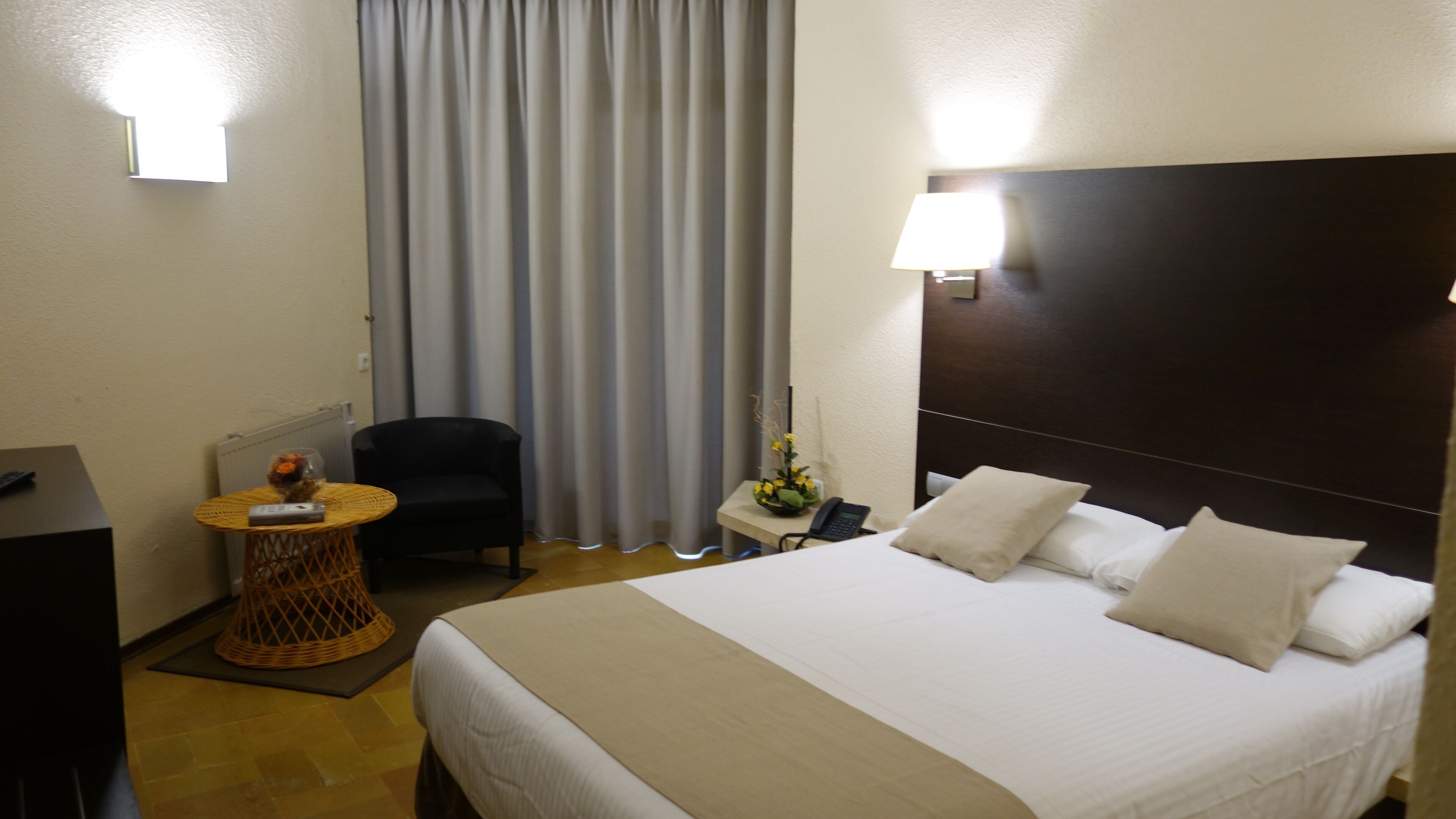 Habitación de hotel con cama, una silla, una mesita con lámpara y ventana con cortinas.