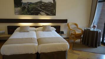 A hotel room with two single beds, a wicker chair, a small table with a vase, and a painting on the wall.
