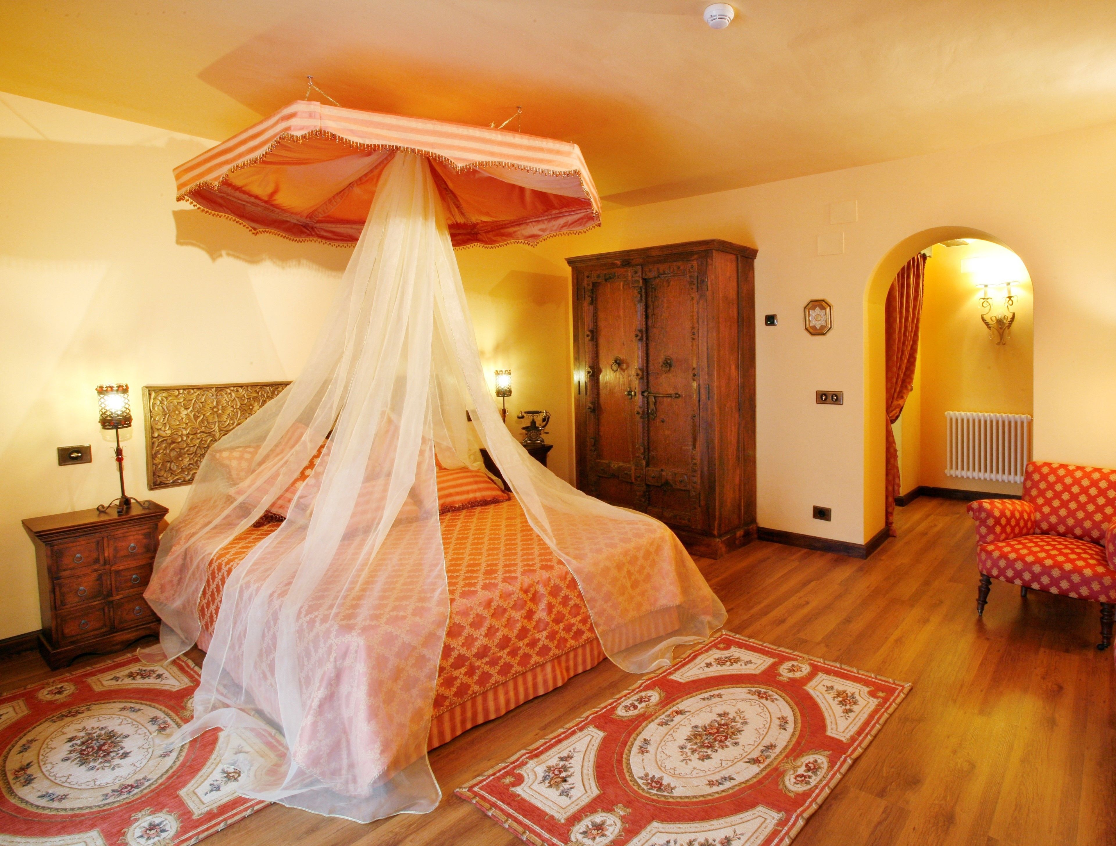 A bedroom with a canopy bed, wooden wardrobe, and a red patterned armchair.