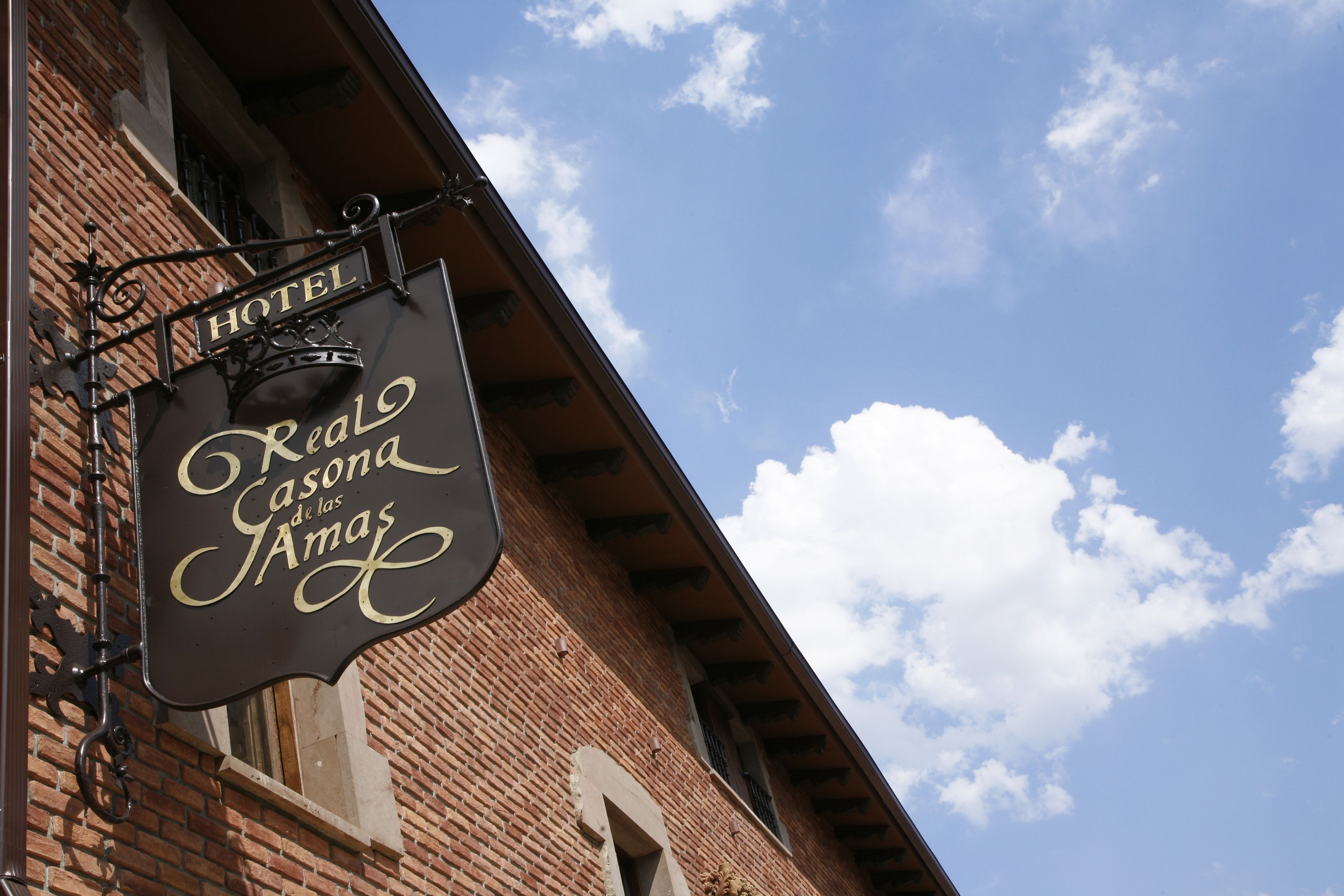 A hotel sign with the name "Real Casa de las Amas" against a brick building under a blue sky with clouds.