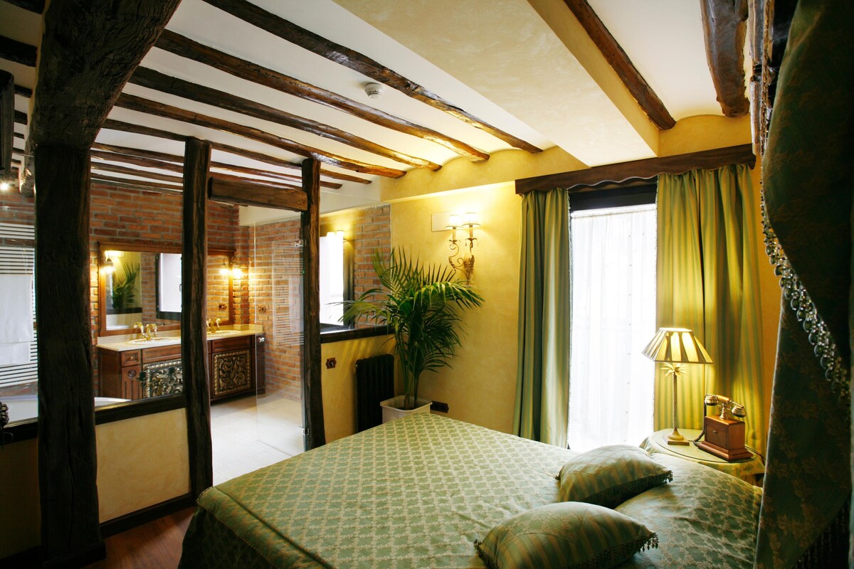 A hotel room with a bed, a nightstand, a lamp, a potted plant, and a view of the bathroom.