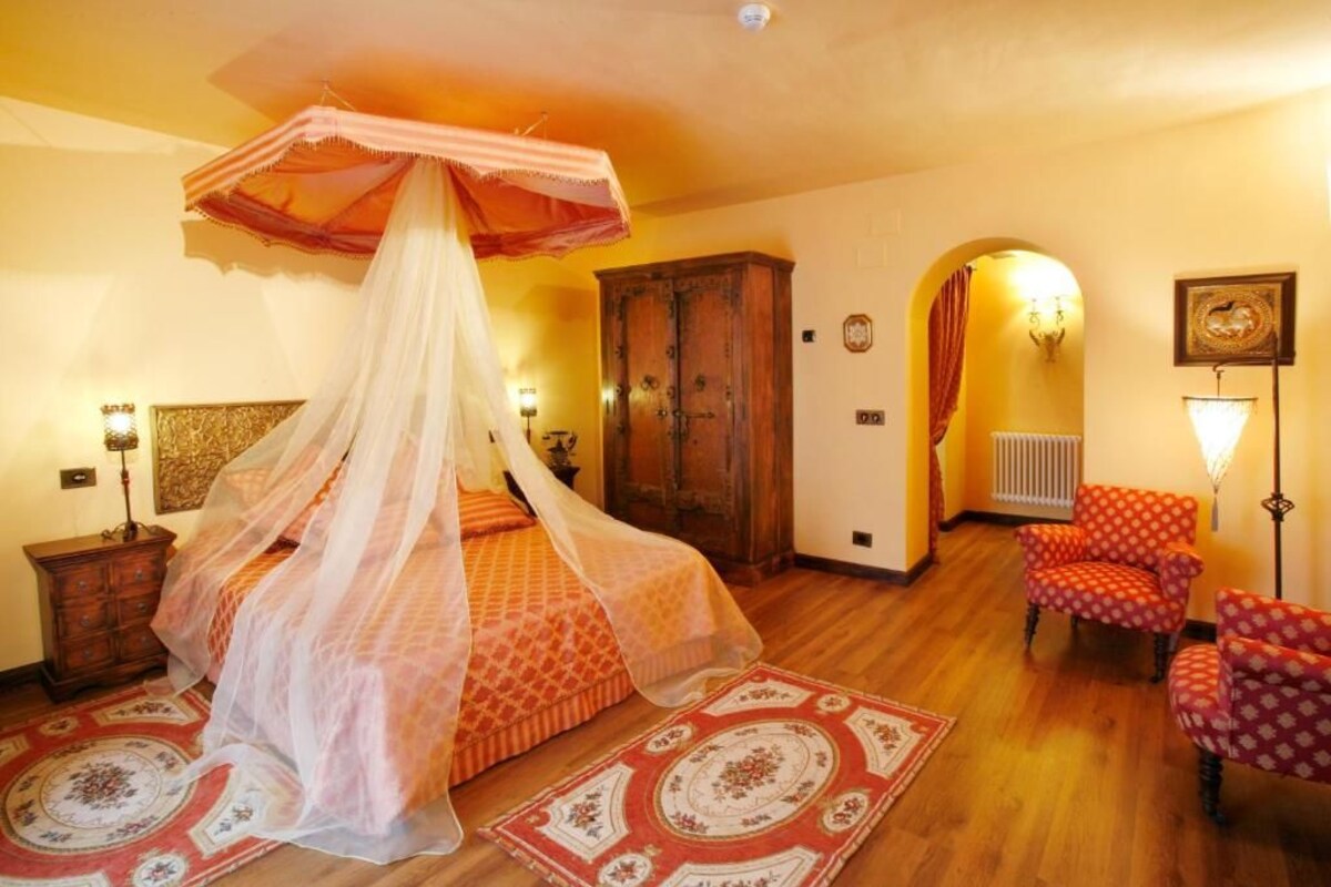A bedroom with a canopy bed, wooden wardrobe, red patterned chairs, and a traditional lamp.