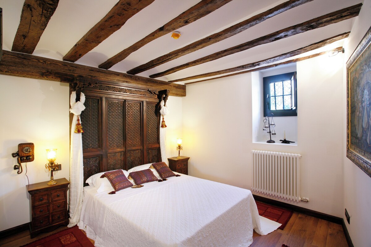 A bedroom with a wooden ceiling, a large bed with white bedding, two bedside tables with lamps, a window with a view, and a radiator.