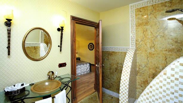 Une salle de bain avec un lavabo en verre, un miroir rond, une porte en bois donnant sur une zone de douche et un mur décoré d’un motif.