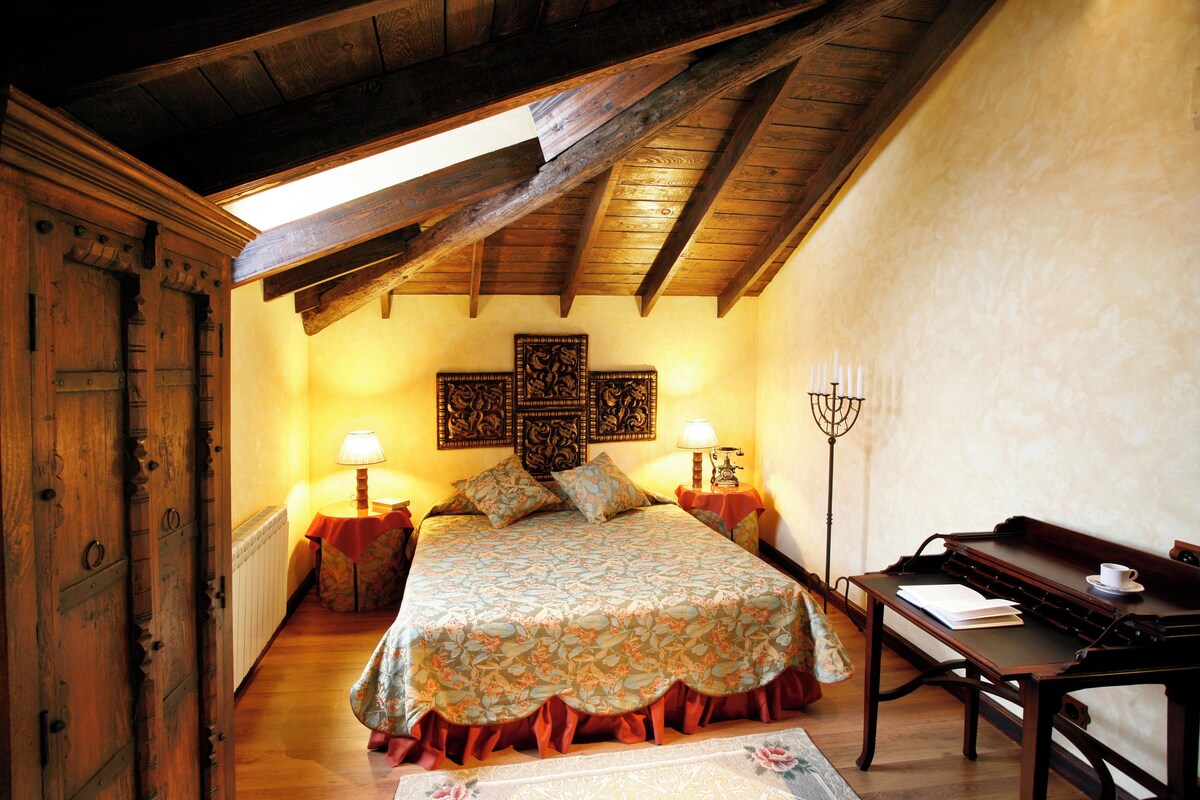 A bedroom with a wooden ceiling, a bed with a floral bedspread, a nightstand with a lamp, and a table with a book and a cup.
