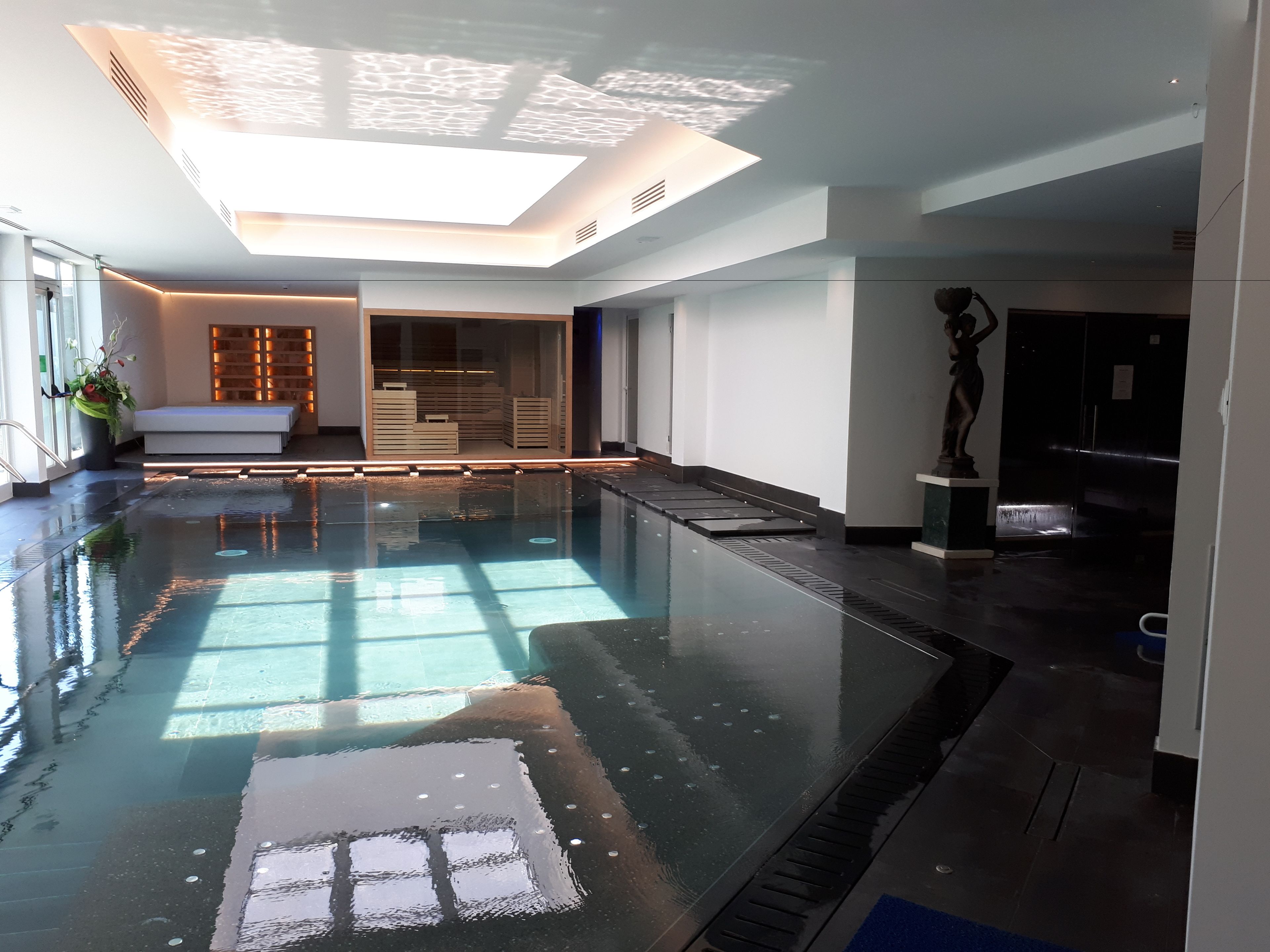 A modern indoor swimming pool with a skylight ceiling, a statue, and a seating area with a plant.