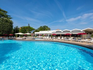 Une piscine entourée de chaises longues et de parasols, elle-même bordée d’un bâtiment doté d’un toit blanc et de parasols rouges.
