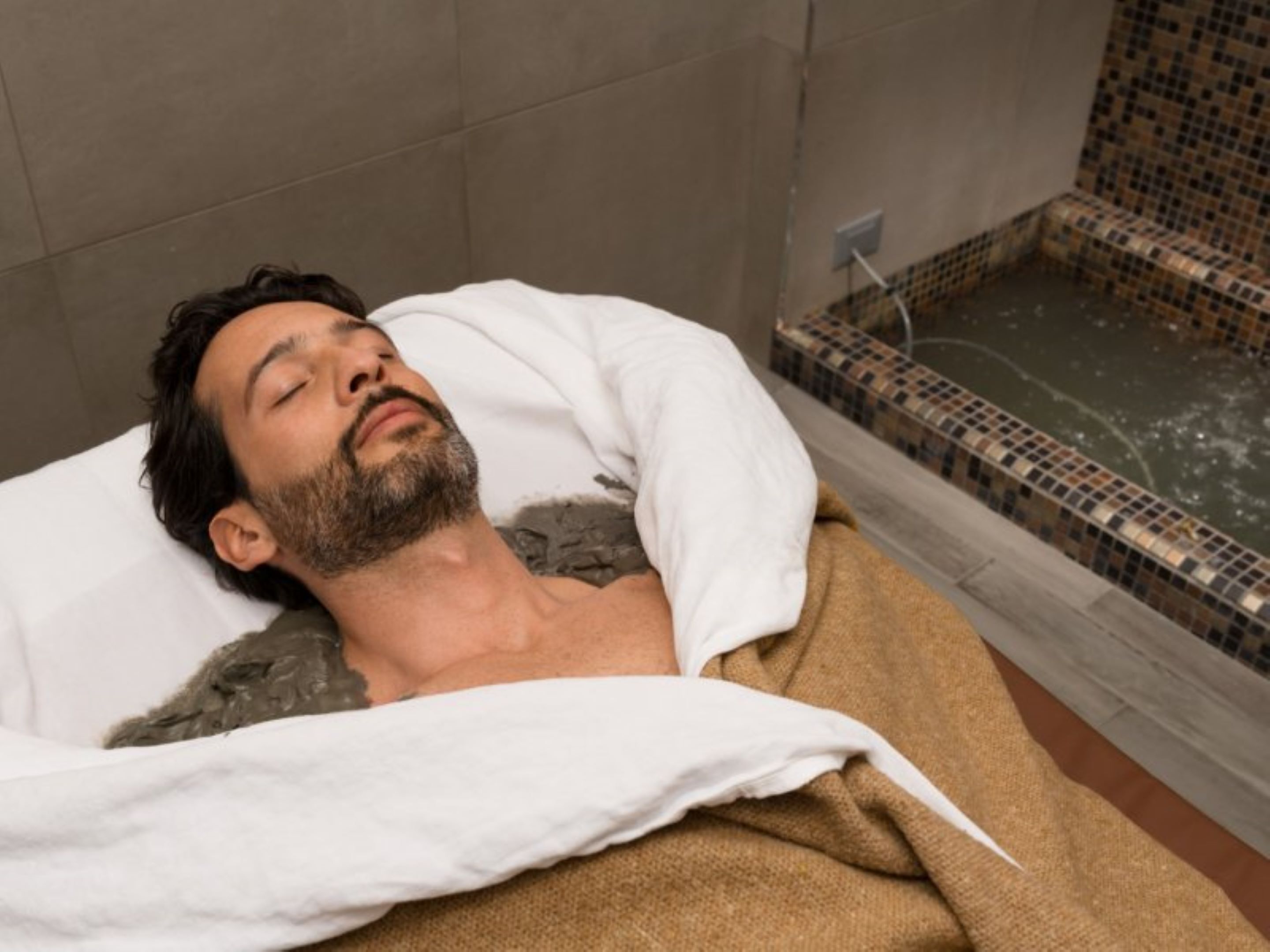 A person lying in a room with a tiled wall and a bathtub.