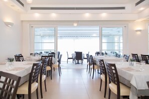 A dining area with multiple tables and chairs, large windows, and a view of the outside. - Hotel Baja  (Arzachena)