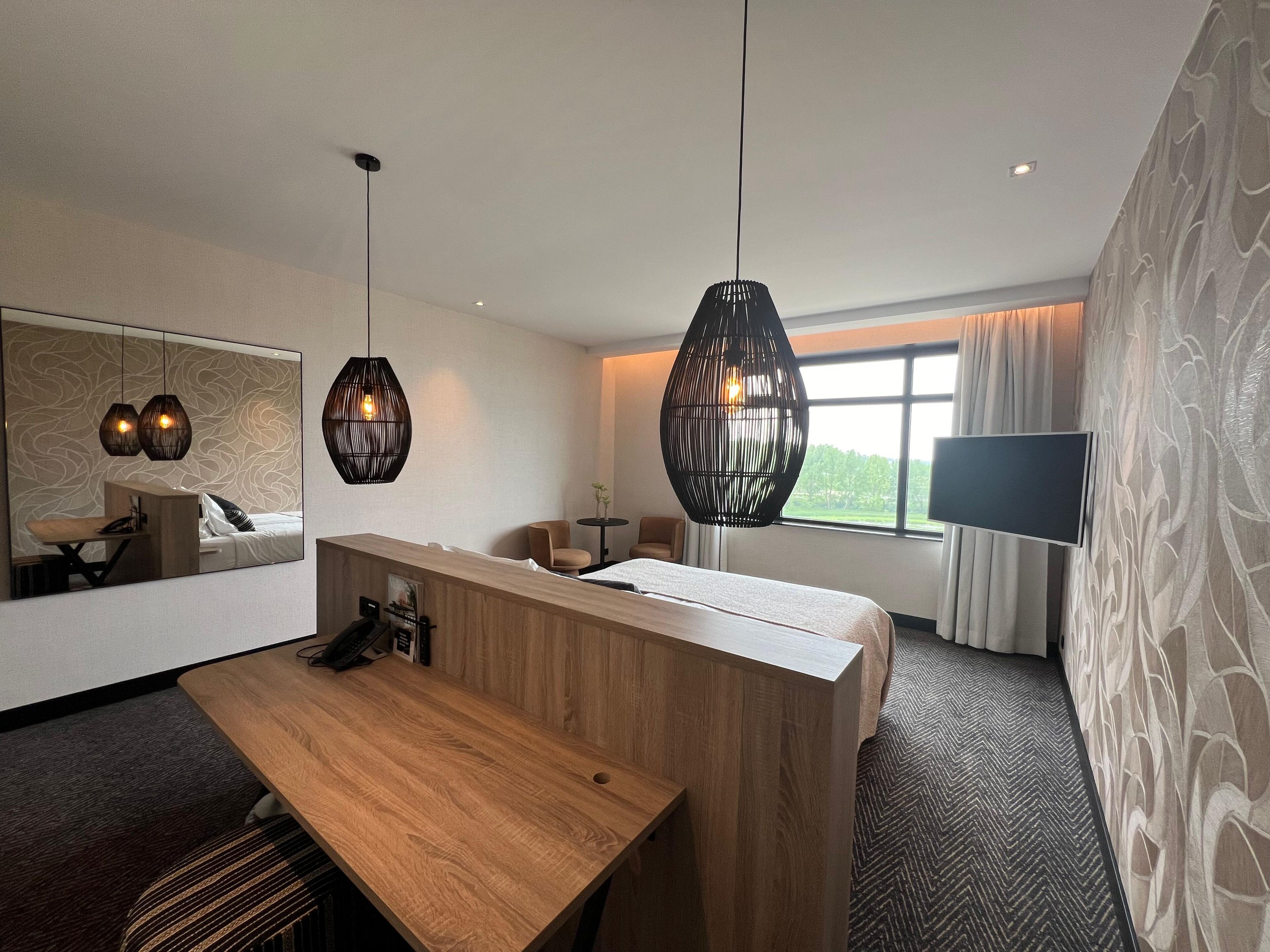 A hotel room with a wooden reception desk, two pendant lights, a large mirror, a flat-screen TV, and a window with curtains.